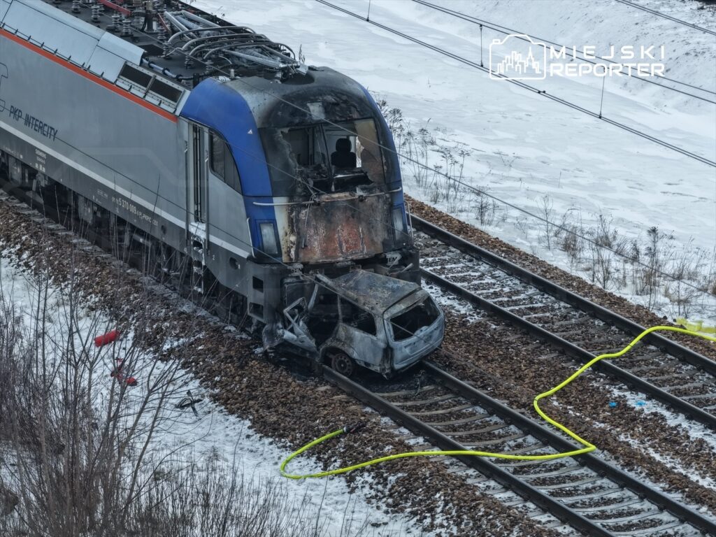 Spalony samochód osobowy leży obok torów kolejowych, obok stoi uszkodzony pociąg w zimowym otoczeniu.