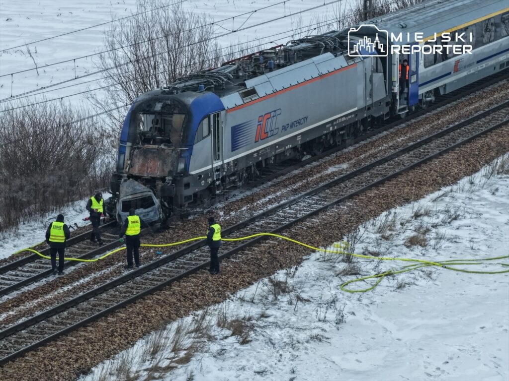 Czterech pracowników w żółtych kamizelkach zabezpiecza miejsce wypadku na torach kolejowych, obok uszkodzonego pociągu.