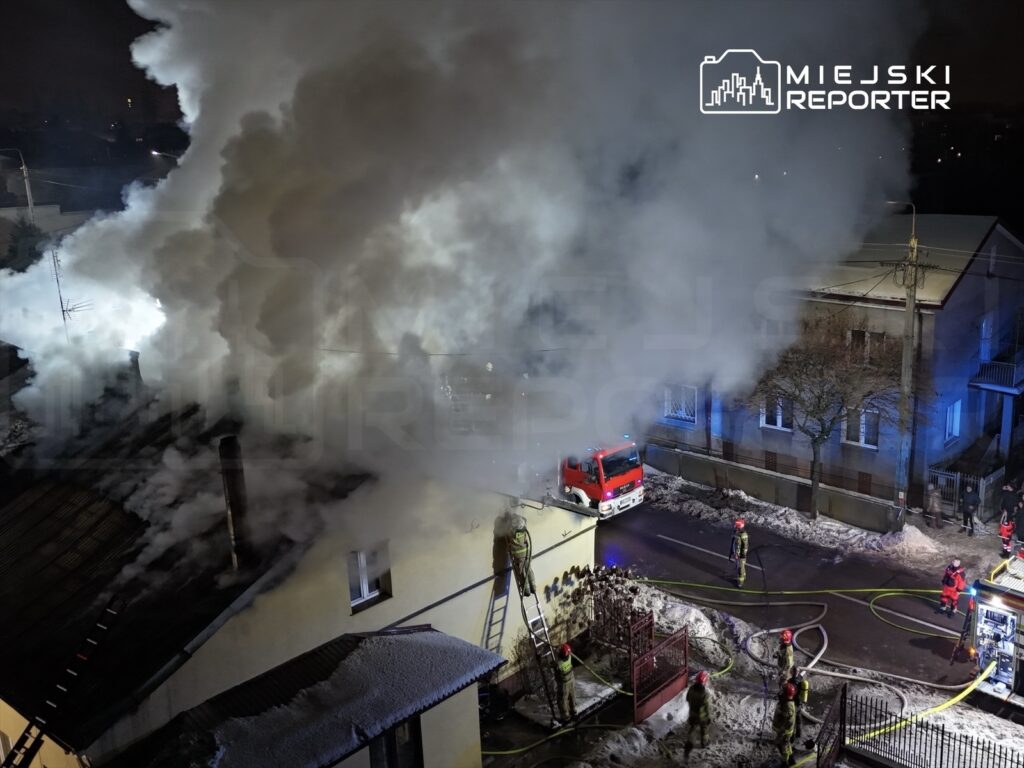Strażacy gaszą pożar w budynku mieszkalnym, otoczeni gęstym dymem i sprzętem gaśniczym.
