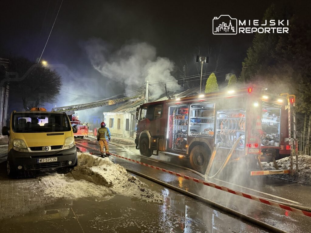 Strażak w mundurze podchodzi do wozu strażackiego na oświetlonej ulicy, gdzie unosi się dym z płonącego budynku.