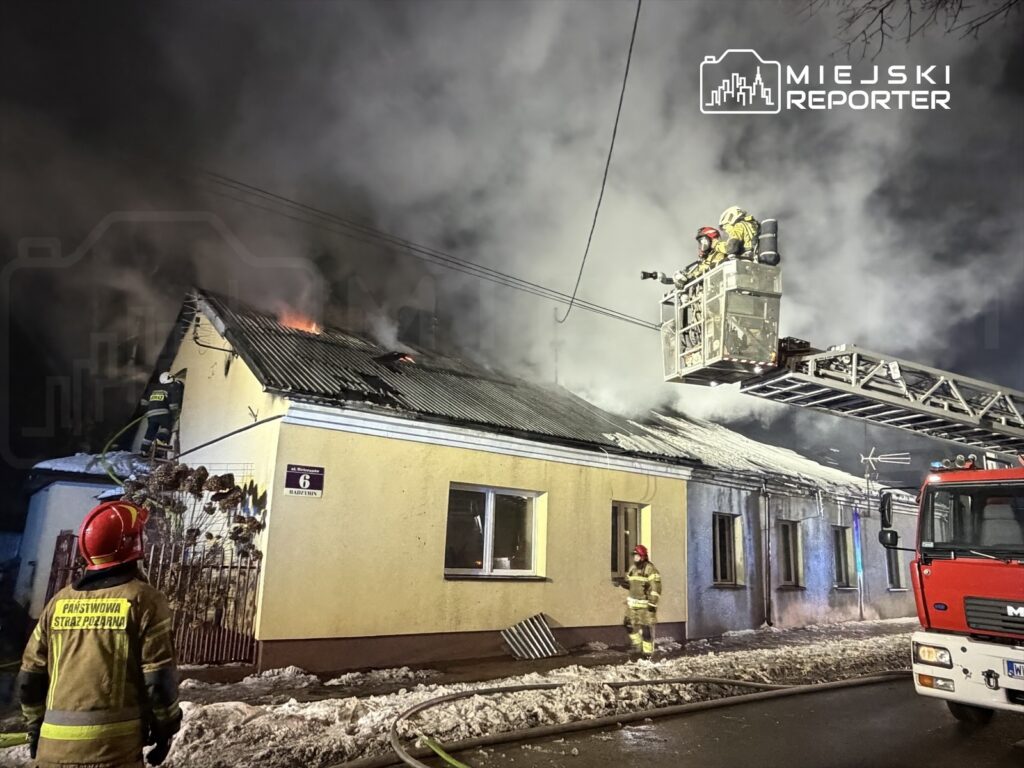 Strażacy gaszą pożar dachu budynku mieszkalnego, używając drabiny mechanicznej w otoczeniu dymu i śniegu.