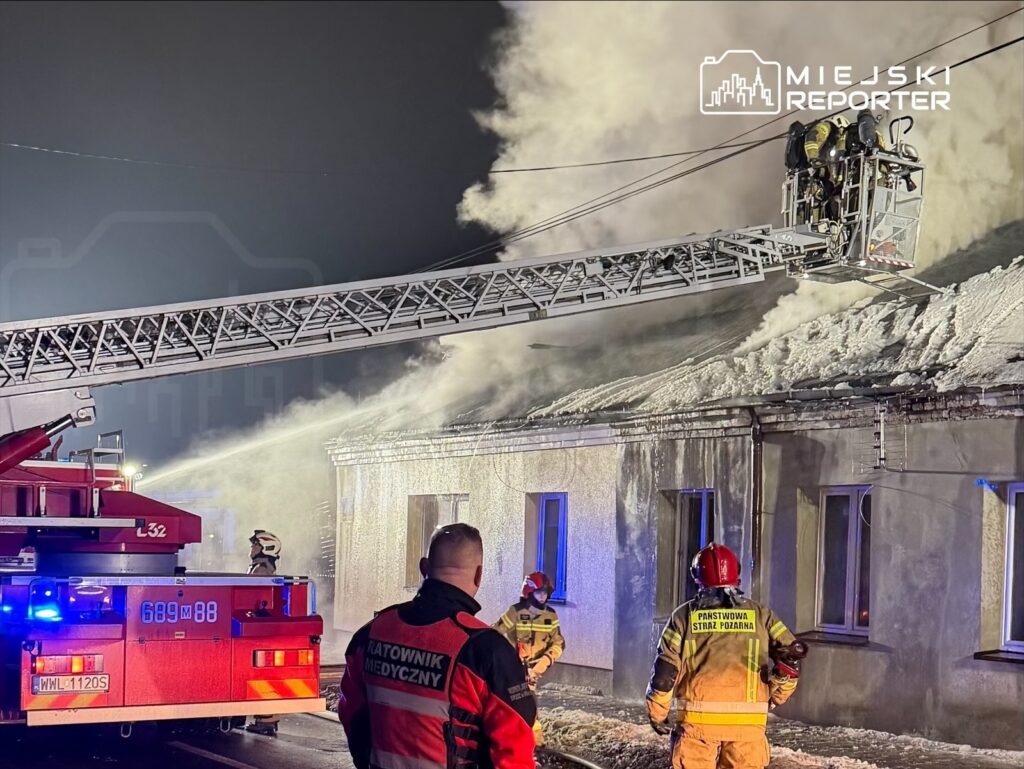 Strażacy na drabinie gaśniczej polewają wodą dach budynku, z którego wydobywa się dym, podczas akcji ratunkowej w nocy.