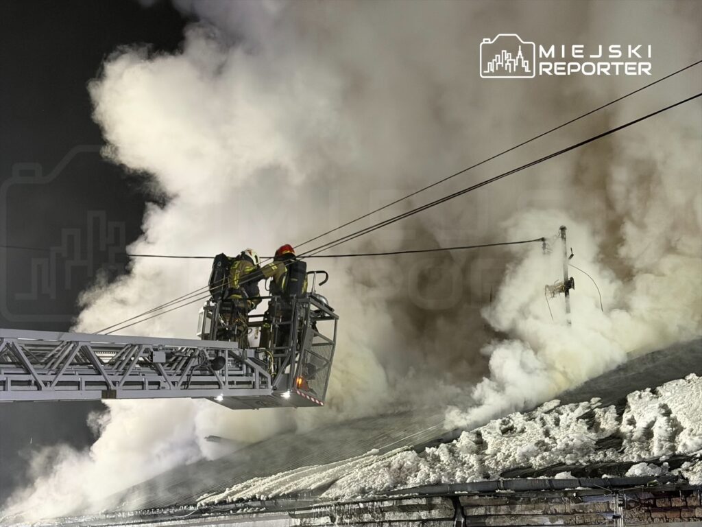 Strażacy na podnośniku gaśniczym polewają dach budynku wodą w trakcie akcji gaśniczej, otoczeni gęstym dymem.