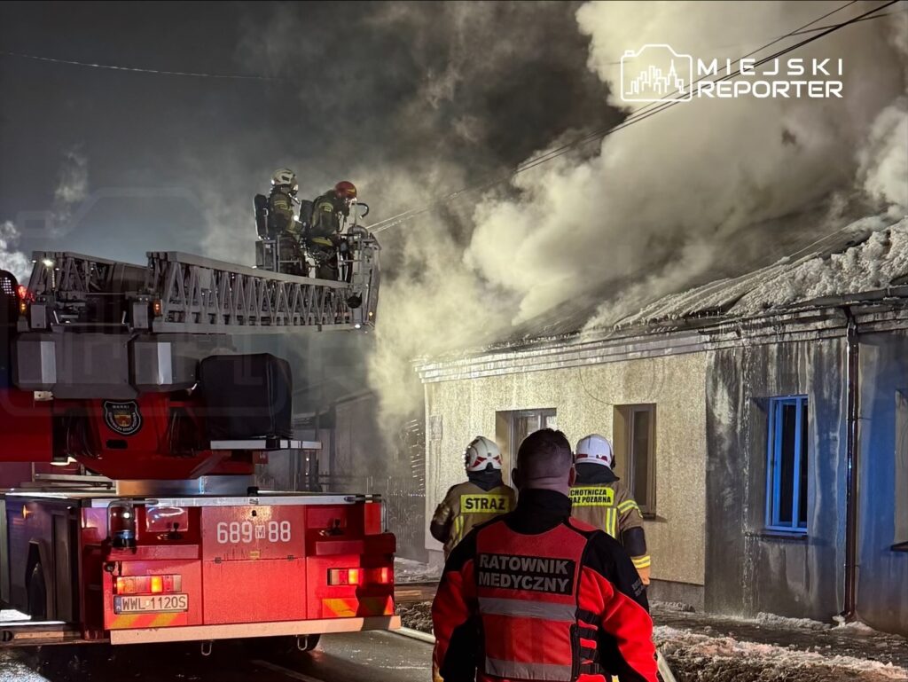Strażacy na podnośniku gaszą pożar dachu budynku, w tle widać gęsty dym i grupę ratowników medycznych.
