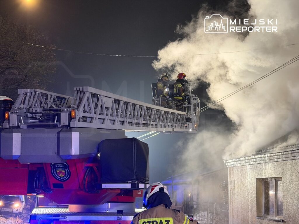 Strażacy w hełmach na podnośniku gaśniczym, gaszący dymiący budynek w nocy.