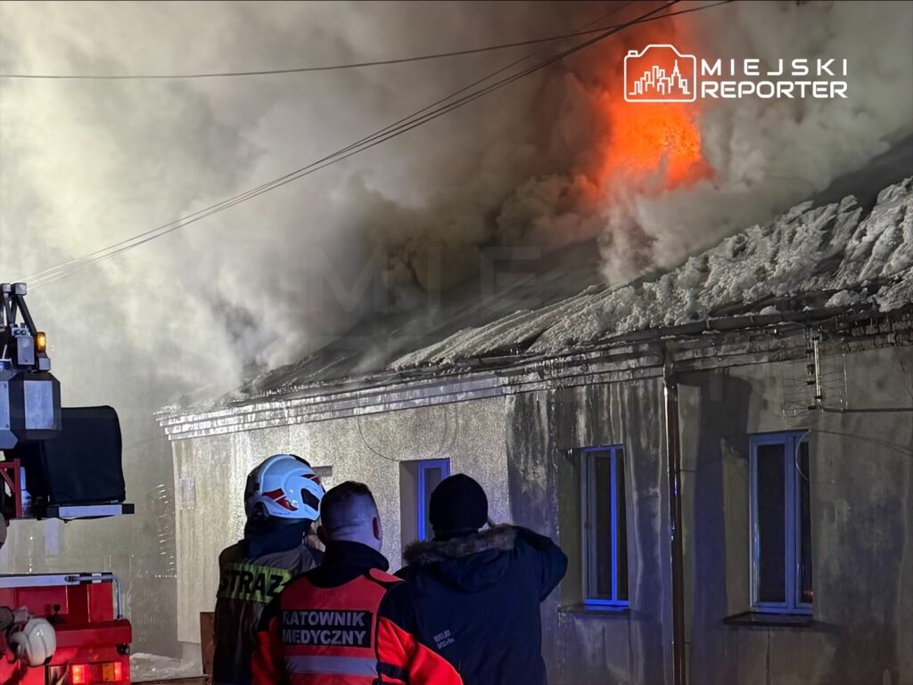 Strażacy i ratownicy medyczni obserwują pożar dachu budynku, z którego wydobywają się gęste kłęby dymu.