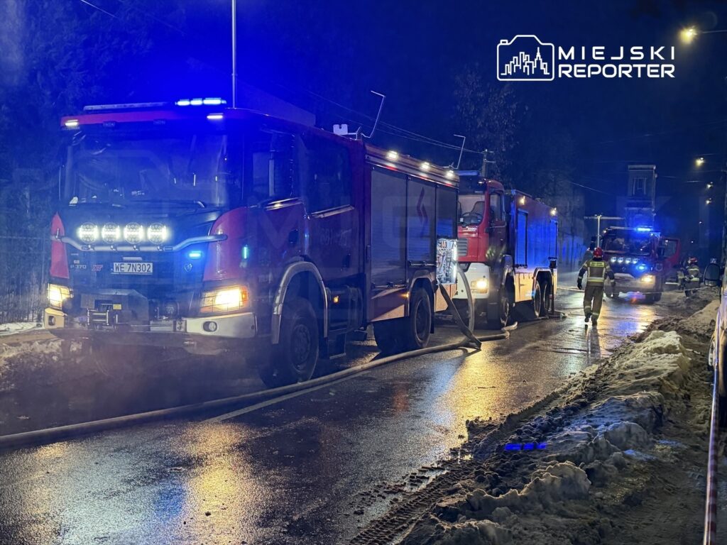 Cztery wozy strażackie na oświetlonej ulicy, strażacy rozkładają węże gaśnicze obok zasypanej śniegiem jezdni.