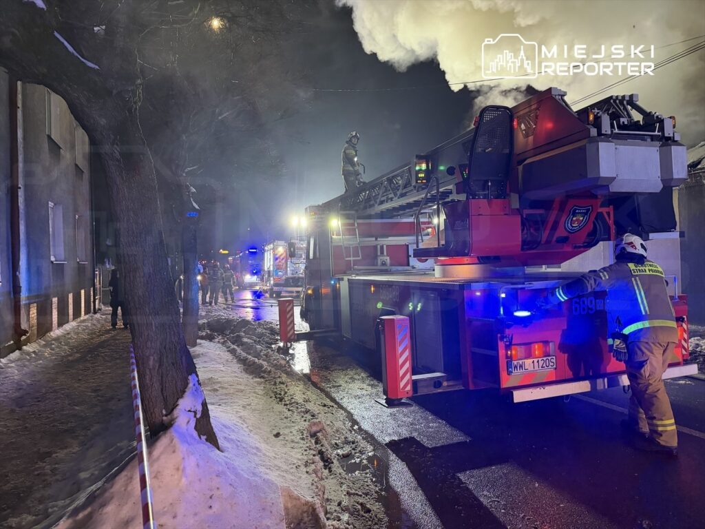 Strażacy przy wozie strażackim w akcji ratunkowej na oświetlonej ulicy, otoczeni przez dym i śnieg.