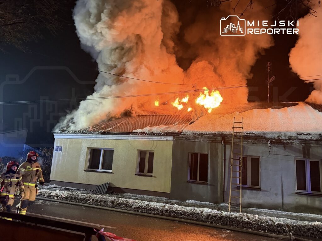 Płonący dach budynku pokryty śniegiem, z dymem i ogniem wydobywającym się z komina. W tle strażacy w mundurach idą do akcji.