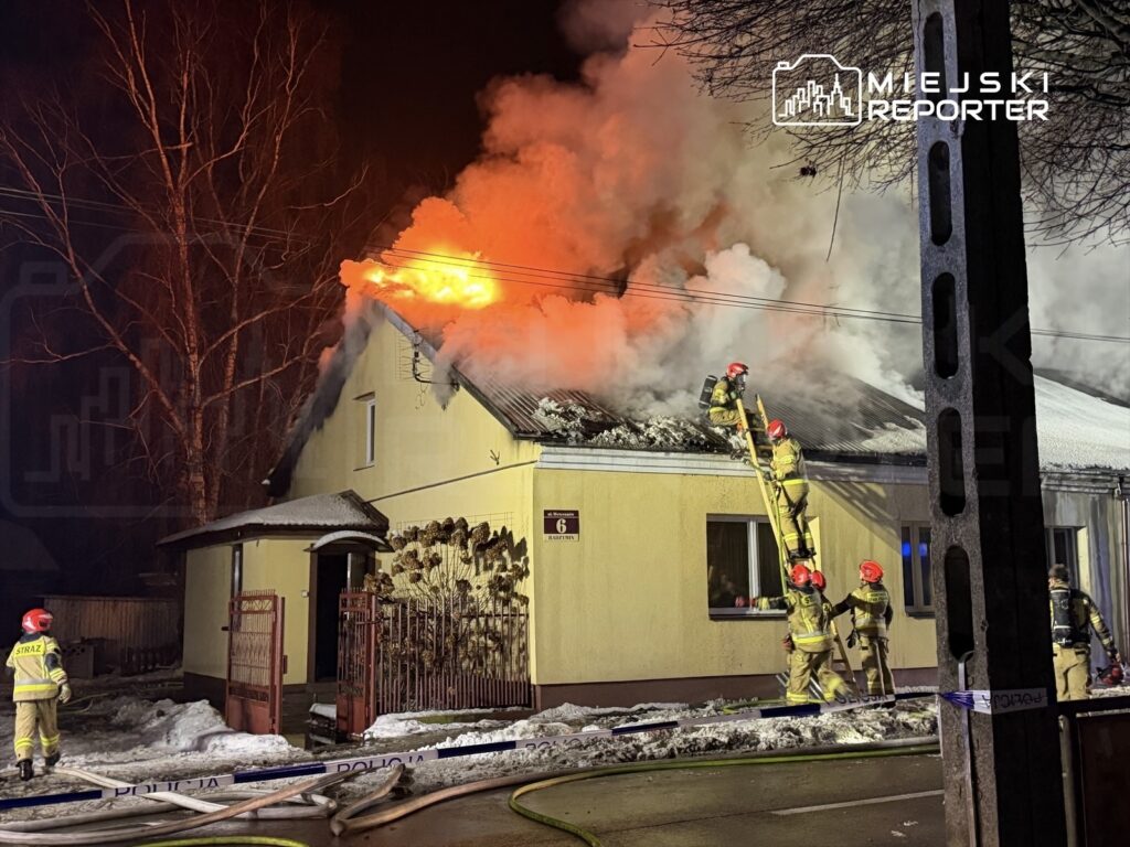 Strażacy gaszą pożar dachu żółtego budynku, w tle widać płomienie i gęsty dym, na ziemi leżą węże gaśnicze.
