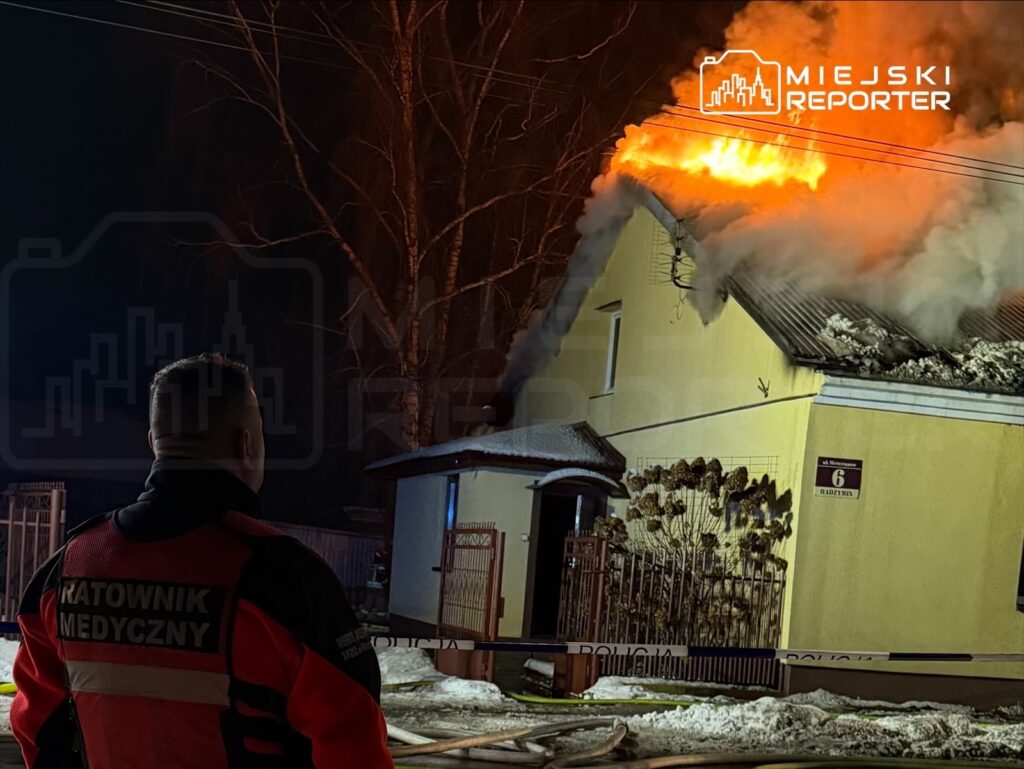 Ratownik medyczny obserwujący pożar dachu żółtego domu w nocy, z płomieniami i dymem wydobywającymi się z budynku.