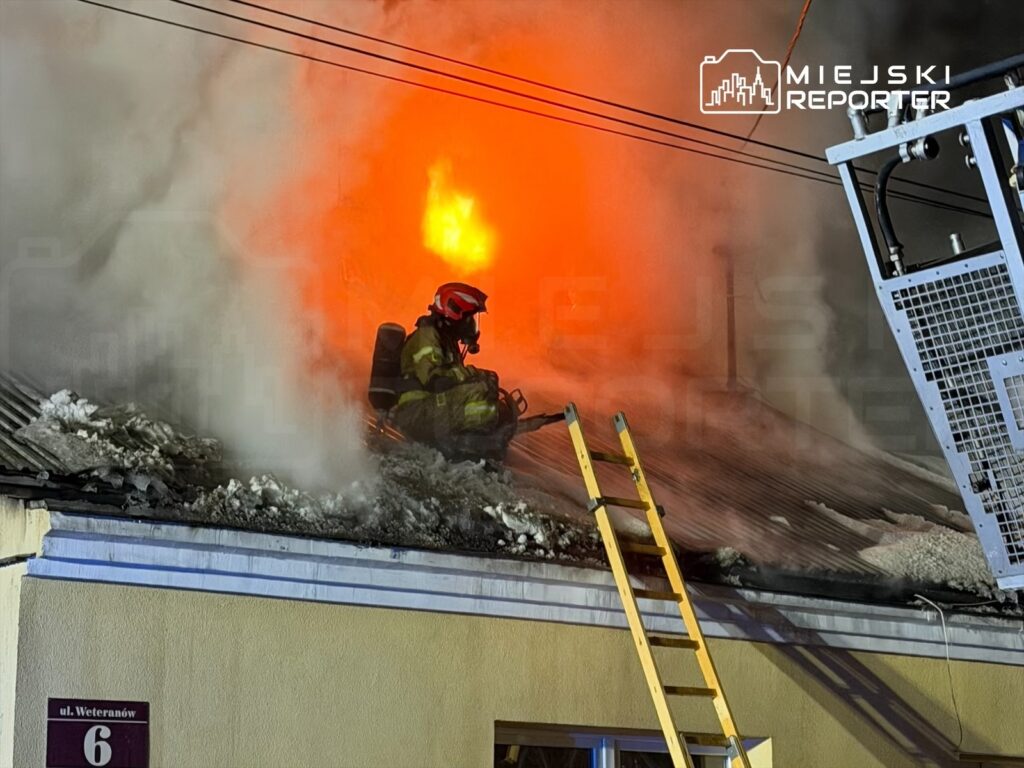 Strażak w hełmie i odzieży ochronnej gasi ogień wydobywający się z dachu budynku, otoczony gęstym dymem.