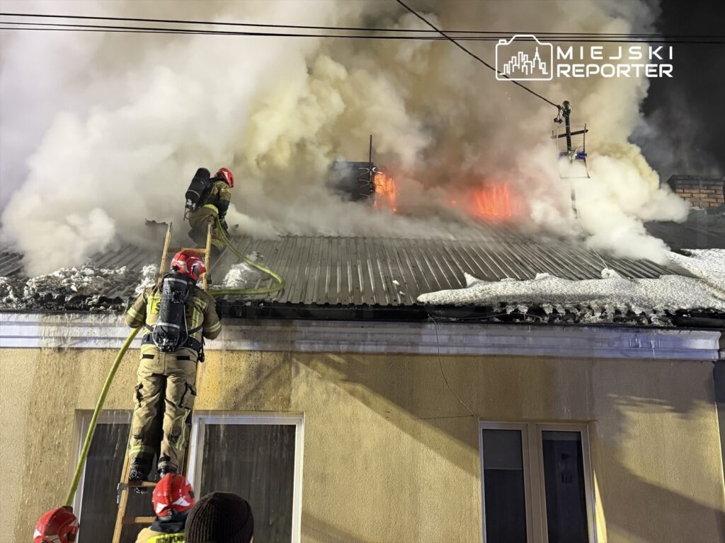 Strażacy na dachu budynku gaszą płonący komin, otoczeni gęstym dymem i ogniem.
