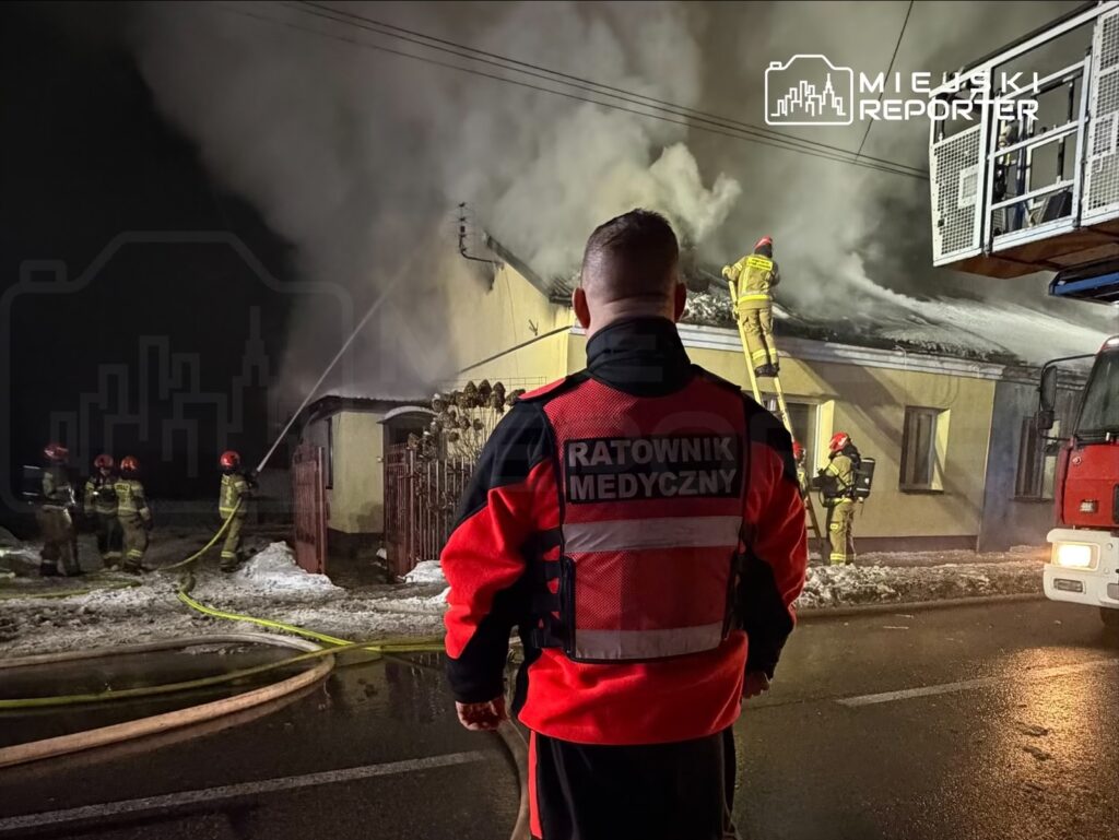 Ratownik medyczny obserwuje akcję gaśniczą, podczas gdy strażacy gaszą pożar na dachu budynku w nocy.