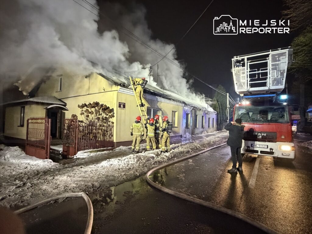 Strażacy w mundurach gaszą pożar dachu budynku, używając drabiny i wozu strażackiego na oświetlonej ulicy.