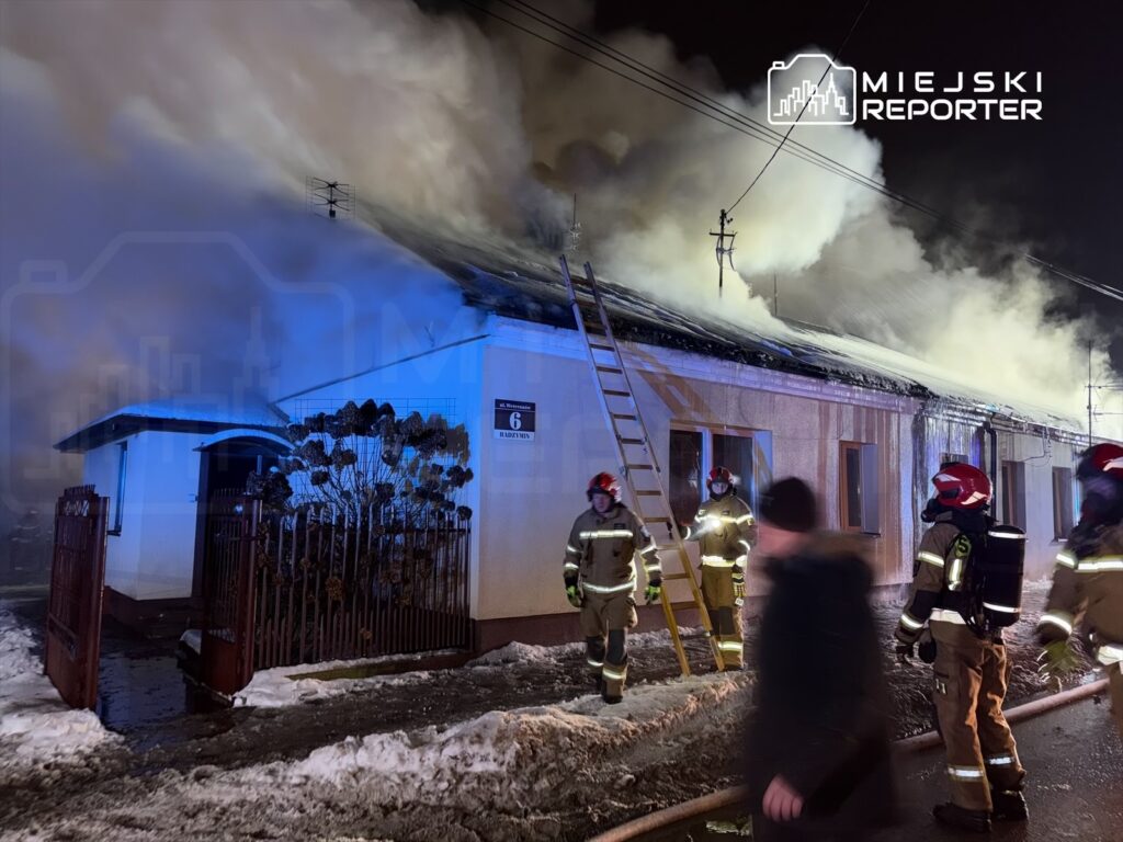 Strażacy w hełmach gaszą pożar dachu budynku mieszkalnego, otoczonego śniegiem i dymem.