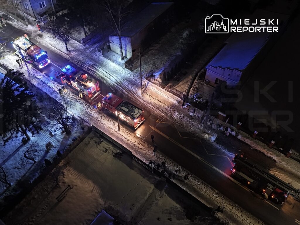 Grupa strażaków pracuje przy pożarze, otoczeni przez kilka wozów strażackich na oświetlonej, zaśnieżonej ulicy.