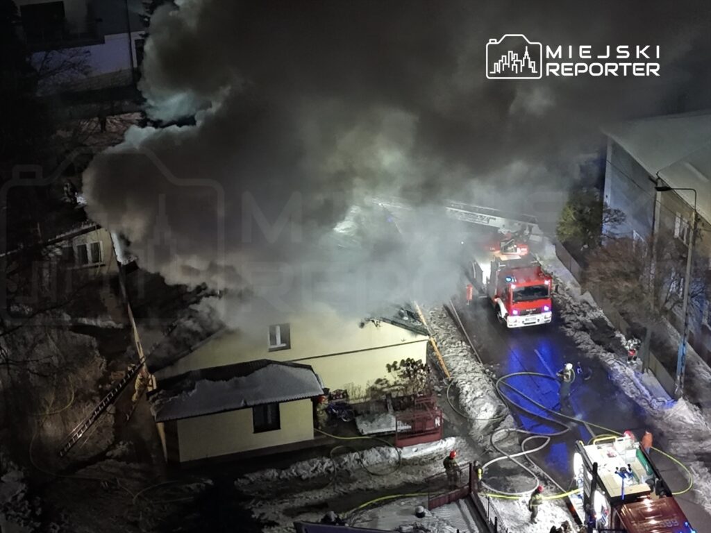 Pożar budynku mieszkalnego, z którego wydobywa się gęsty dym. Strażacy pracują przy wozie strażackim na oświetlonej ulicy.