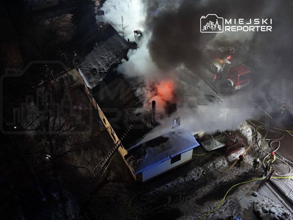 Pożar budynku mieszkalnego, z którego wydobywa się dym, a w tle strażacy gaszą ogień.