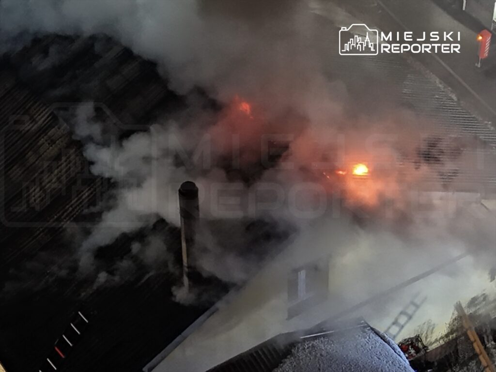 Ogień i gęsty dym wydobywają się z dachu budynku, w tle widoczna komin i fragmenty konstrukcji.