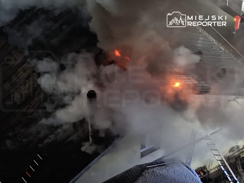 Ogień wydobywający się z dachu budynku, otoczony gęstym dymem, z widoczną drabiną strażacką w tle.