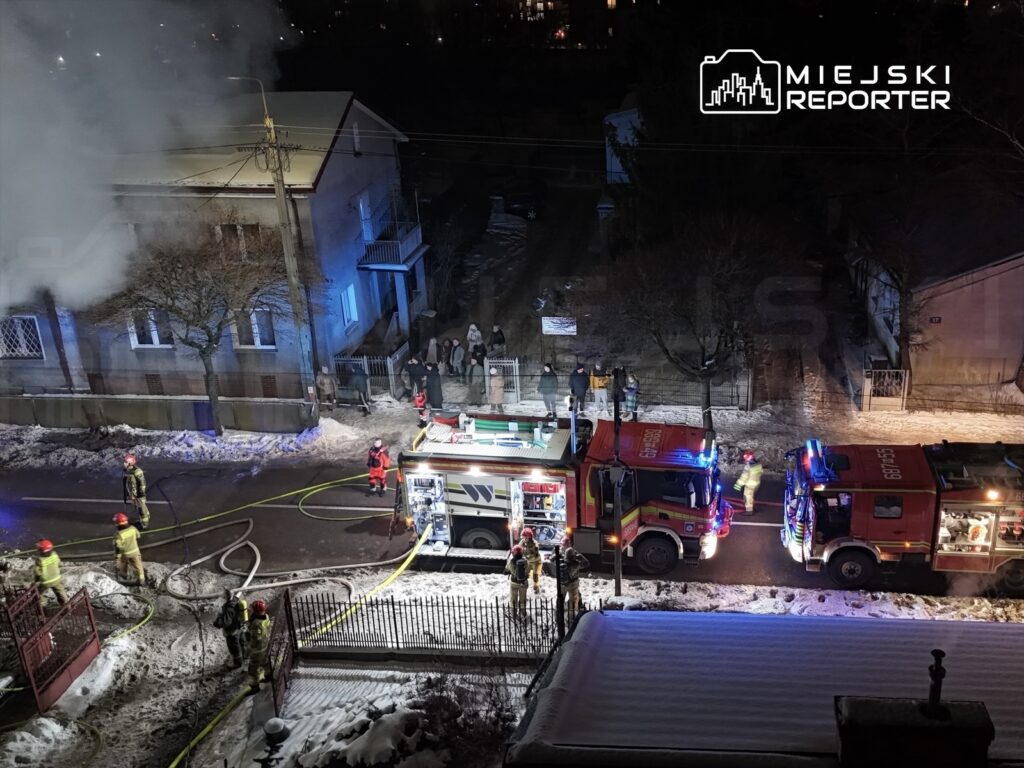 Strażacy w hełmach gaszą pożar budynku, obok stoją wozy strażackie, a w tle widać zebranych ludzi.