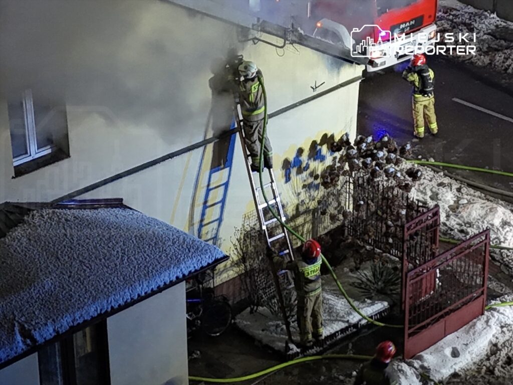 Strażacy na drabinie gaszą pożar na ścianie budynku, a inni członkowie ekipy prowadzą działania w pobliżu.