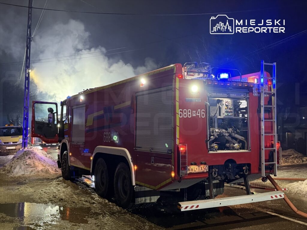 Czerwony wóz strażacki z otwartymi drzwiami, oświetlony niebieskimi i czerwonymi lampami, w otoczeniu dymu.