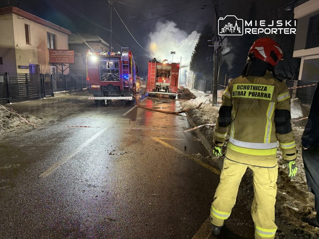 Strażacy w mundurach gaszą pożar w budynku przy ulicy, obok stoją wozy strażackie z włączonymi sygnałami.