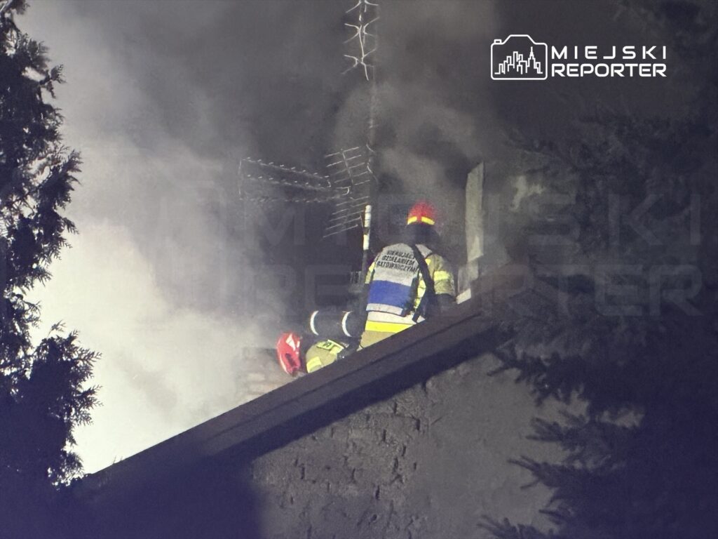 Strażak w hełmie i mundurze przeszukuje dach budynku w gęstym dymie, walcząc z pożarem.