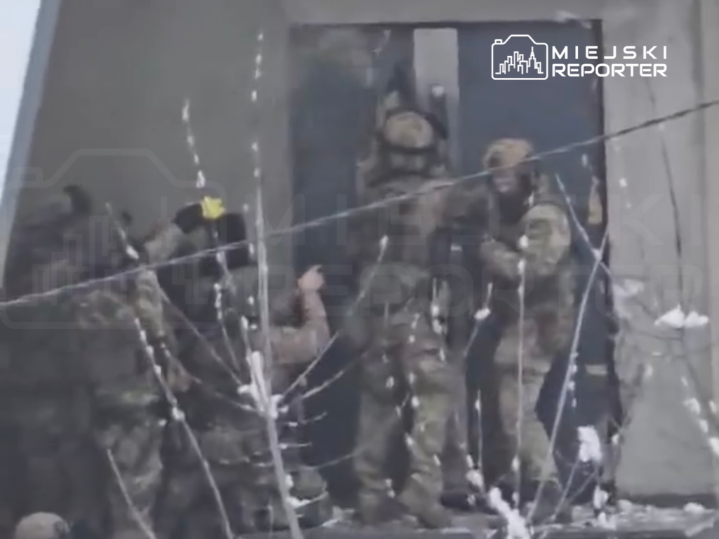 Grupa żołnierzy w kamuflażu wspiera się nawzajem podczas akcji w zniszczonym budynku, otoczonym śniegiem.