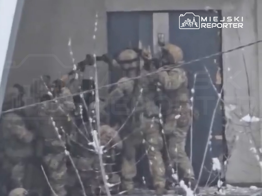 Grupa żołnierzy w mundurach i hełmach przygotowuje się do wejścia do budynku z zamkniętymi drzwiami.