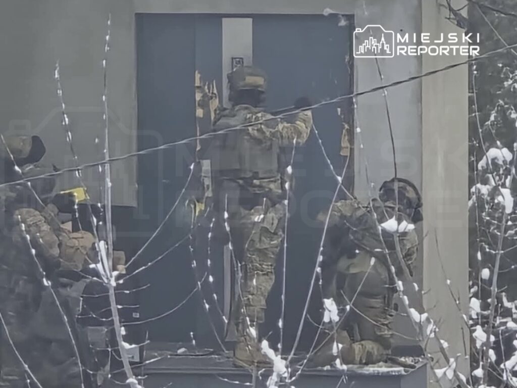 Grupa uzbrojonych żołnierzy w mundurach zbliża się do drzwi budynku, otoczonych drutem kolczastym.