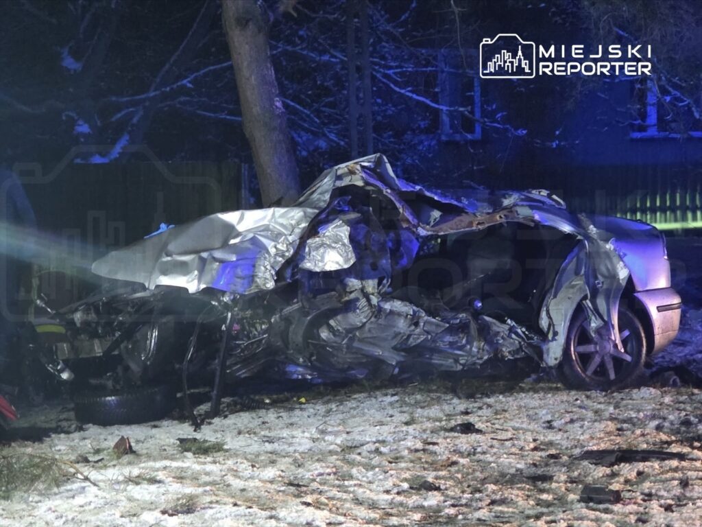 Zniszczony samochód osobowy leży na poboczu drogi, otoczony przez śnieg i oświetlony niebieskimi światłami policyjnymi.