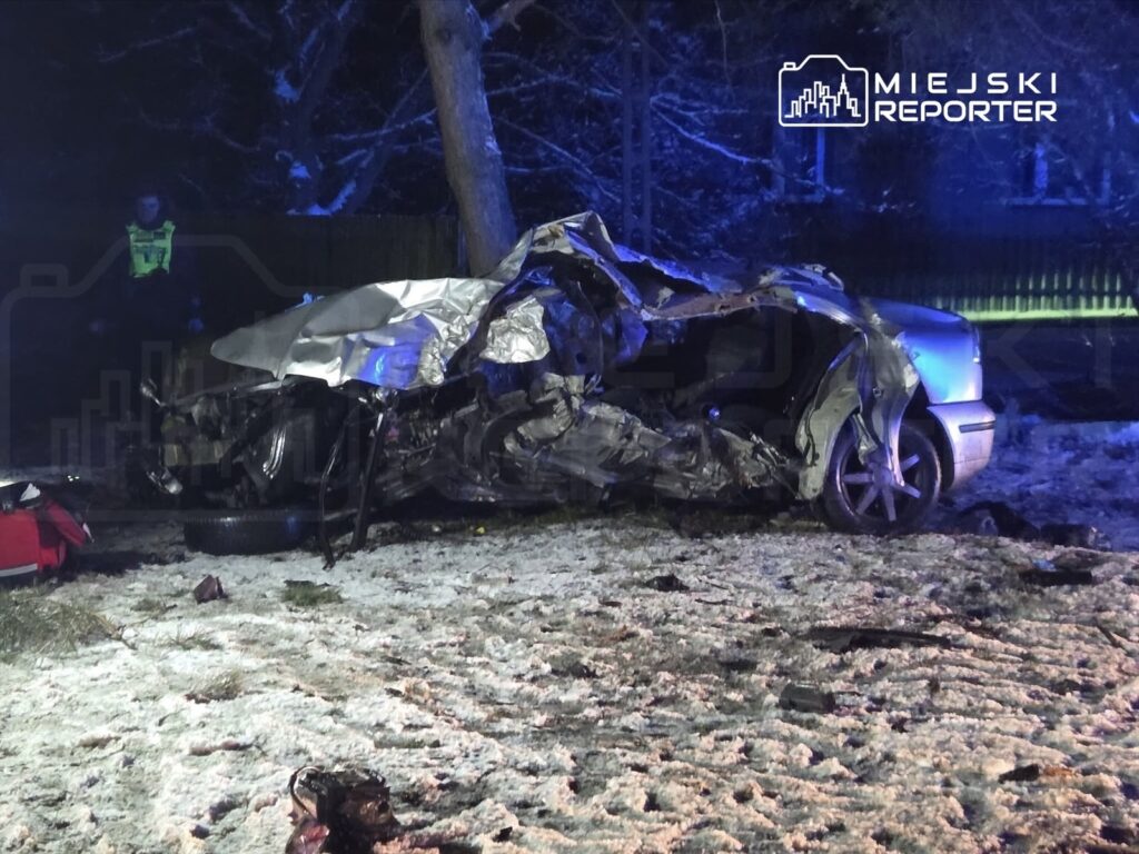 Uszkodzony samochód leżący na poboczu drogi, obok niego ratownik medyczny w akcji, w tle nocna sceneria z oświetleniem policyjnym.