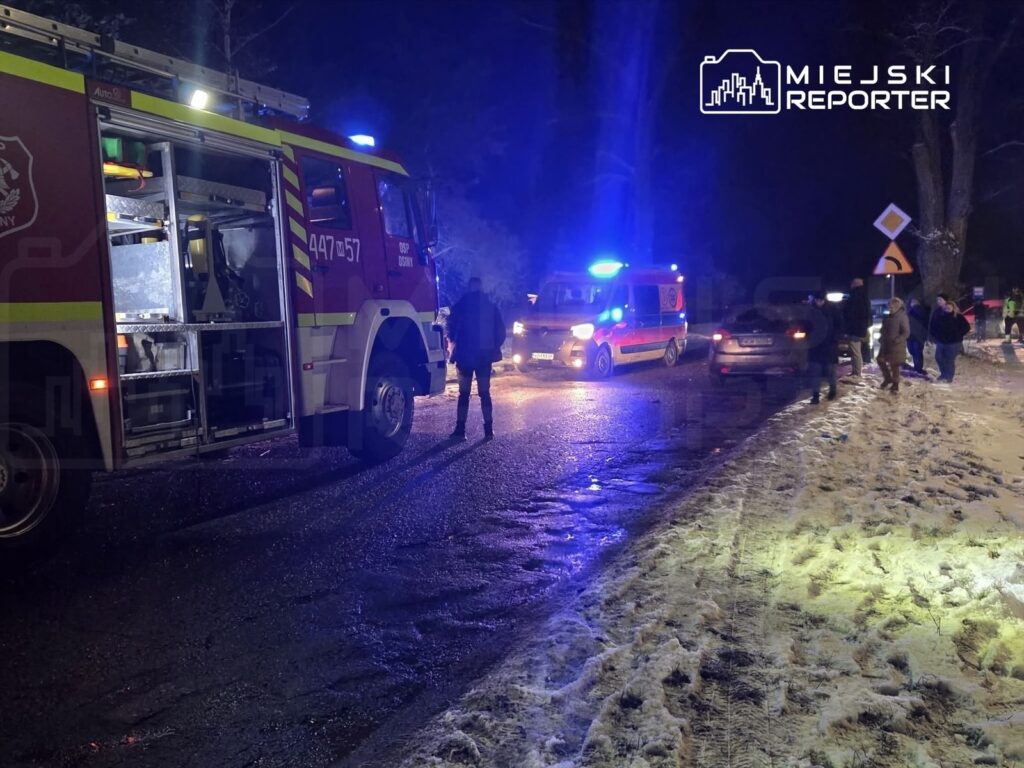 Strażacy przy wozie strażackim interweniują na oświetlonej drodze, obok zatrzymane pojazdy i osoby w zimowym otoczeniu.
