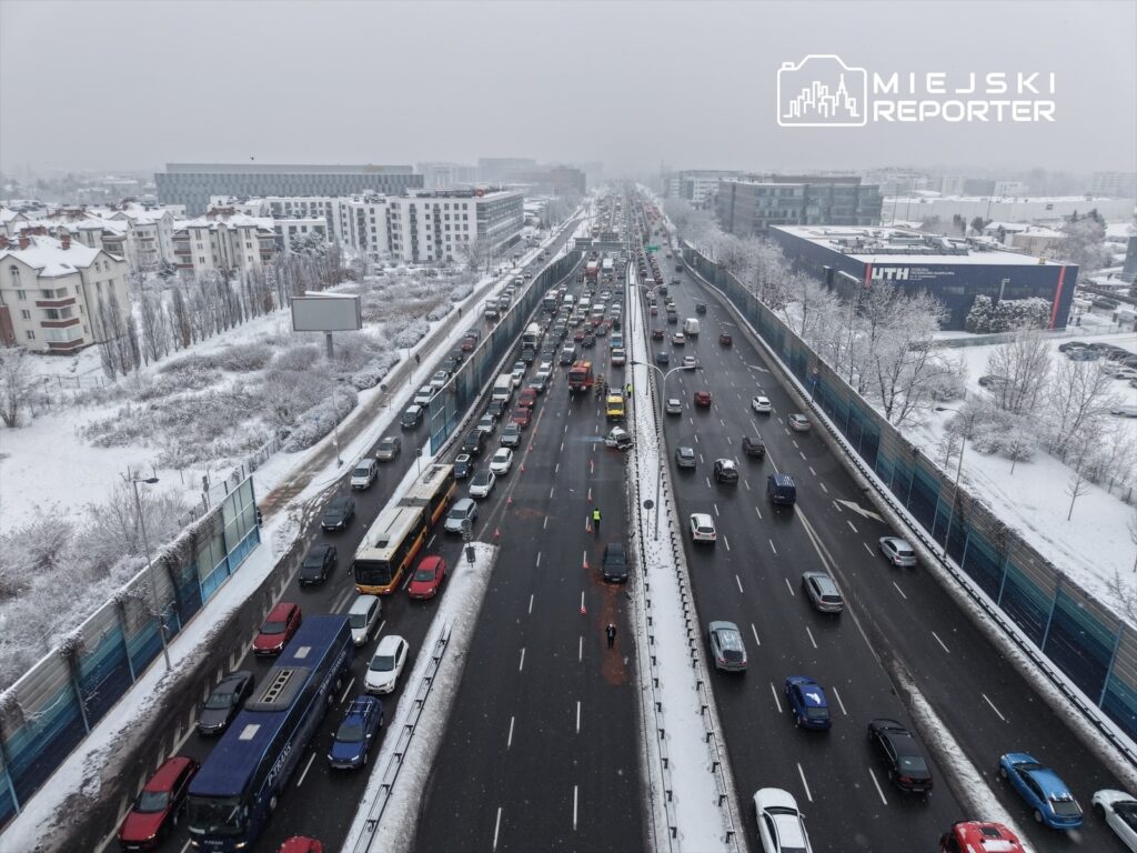 Zatłoczona droga w zimowej scenerii, samochody stoją w korku, a w tle widać nowoczesne budynki i śnieg.