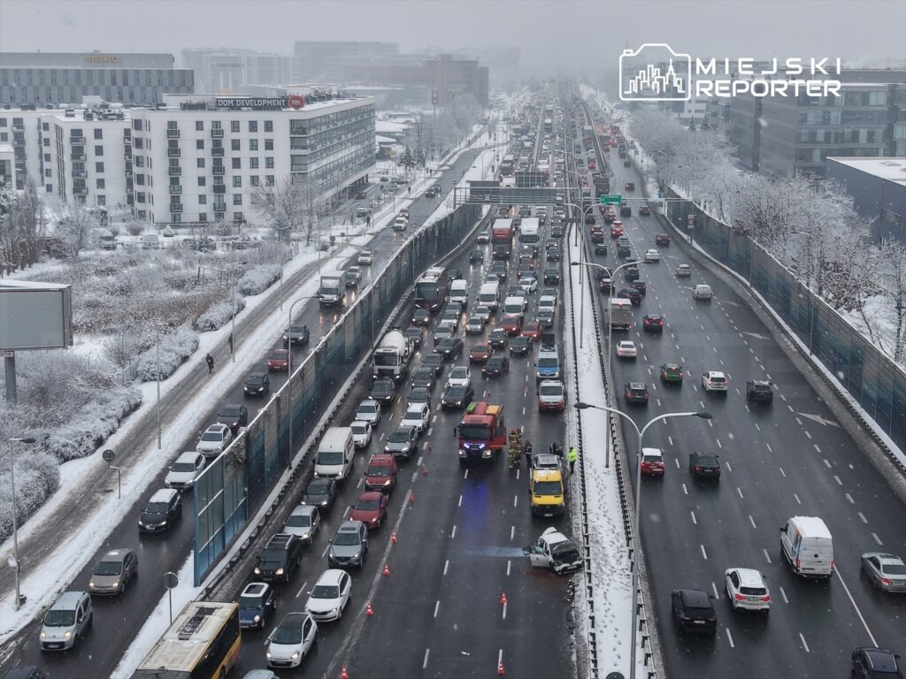 Ruch samochodowy zatrzymany na zaśnieżonej drodze, z wozem strażackim i ekipą ratunkową przy uszkodzonym pojeździe.