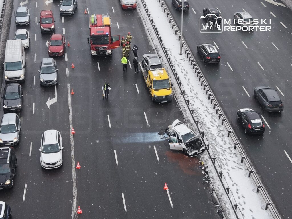 Na drodze widać wypadek samochodowy, z uszkodzonym pojazdem i rozlaną cieczą. Obok interweniują strażacy oraz policja.