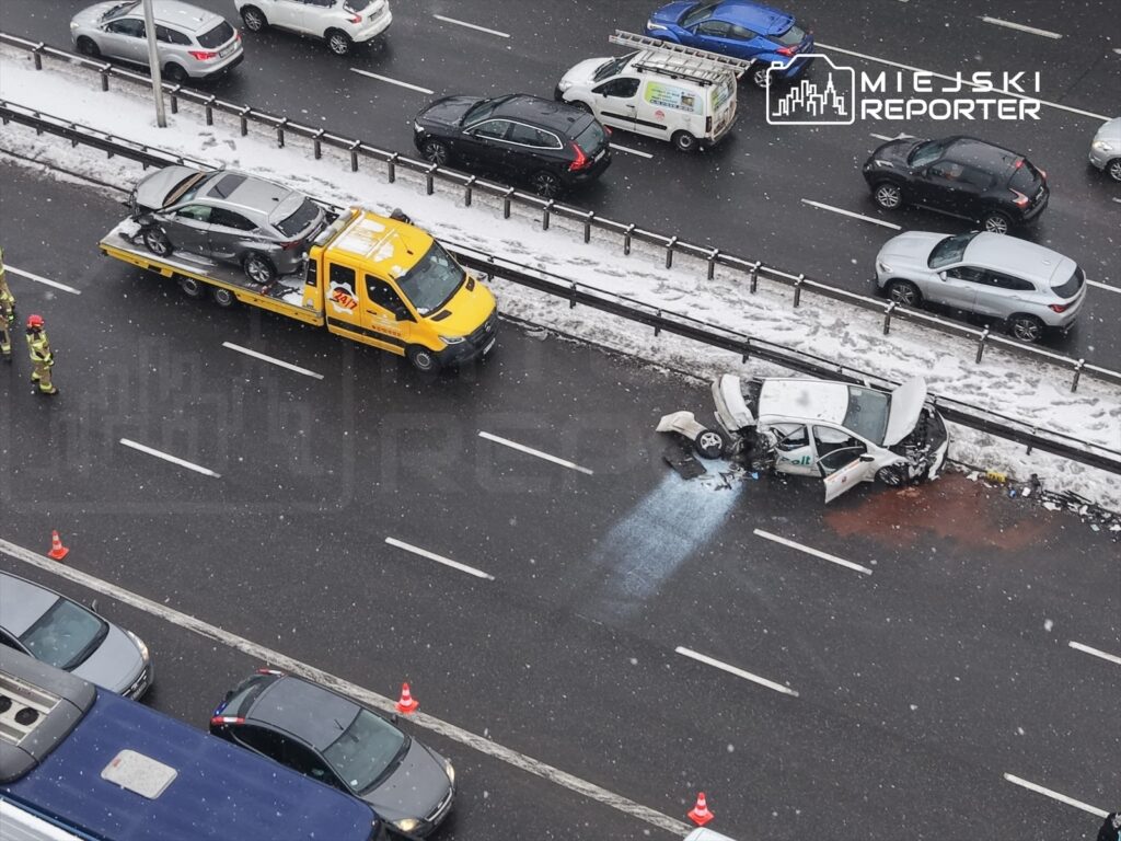 Wypadek drogowy na autostradzie, uszkodzony samochód osobowy z otwartą maską, obok ciężarówka holownicza oraz strażacy.