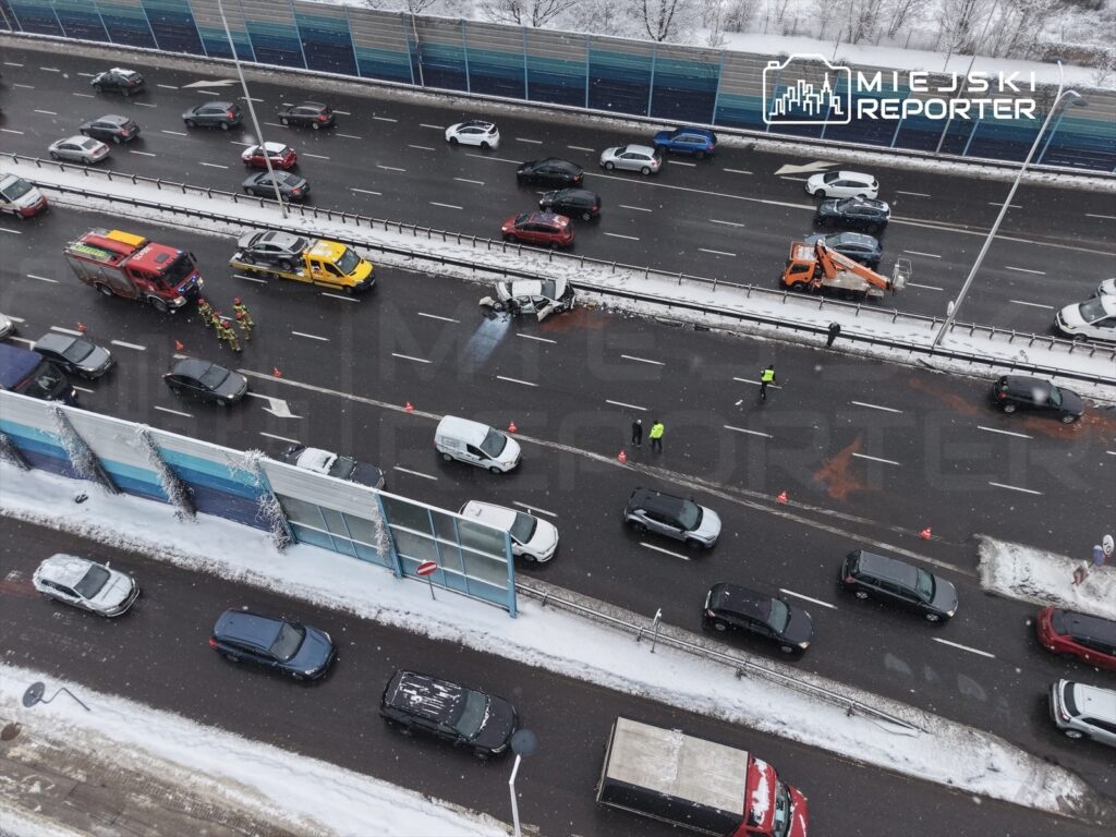Na ruchliwej drodze widać wypadek samochodowy; strażacy i policjanci zabezpieczają miejsce zdarzenia.