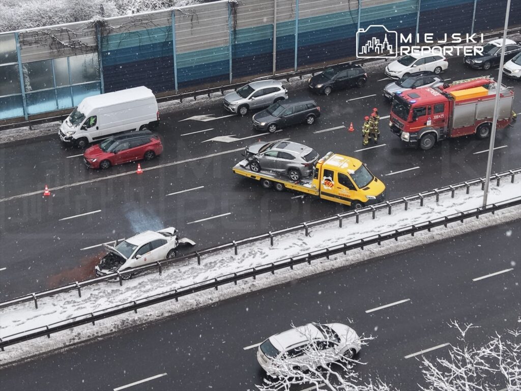 Zespół strażaków pracuje przy wypadku drogowym na zaśnieżonej jezdni, obok stoi laweta z uszkodzonym pojazdem.