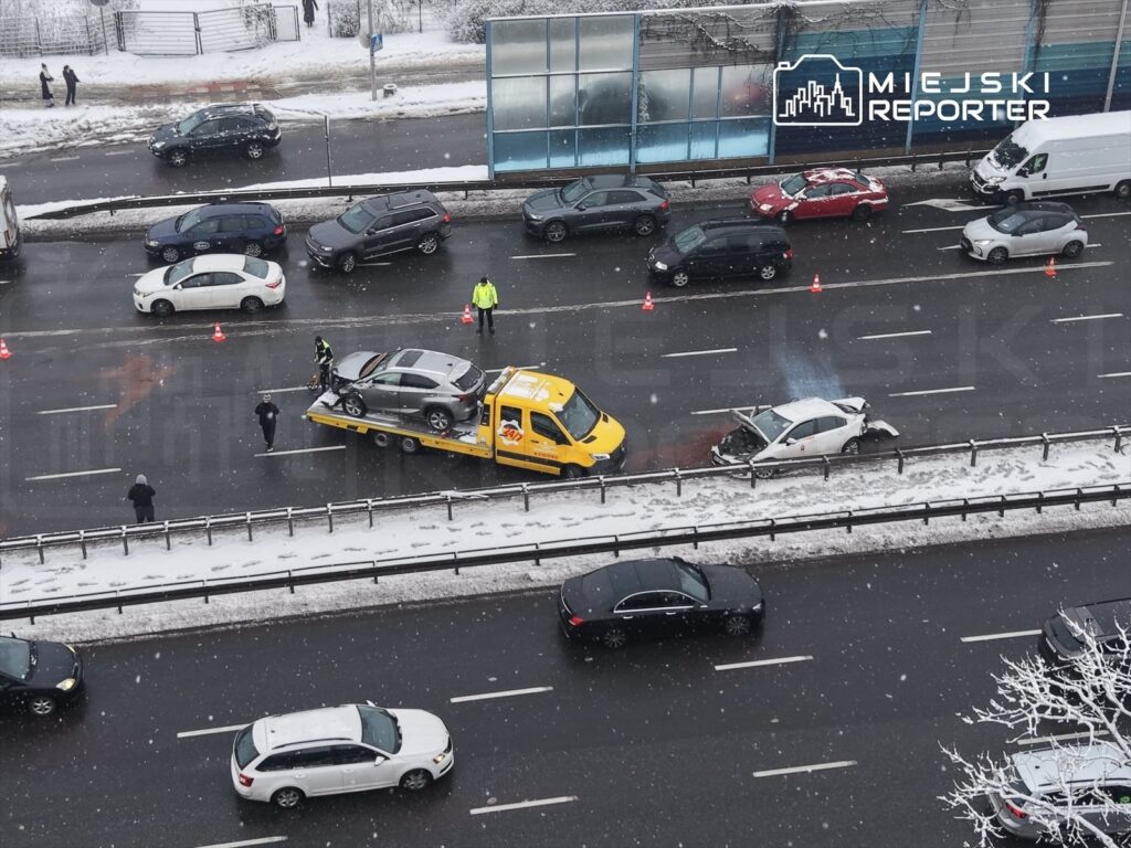 Na drodze zaśnieżonej widać wypadek samochodowy oraz holownik podnoszący uszkodzony pojazd. Ruch jest spowolniony.