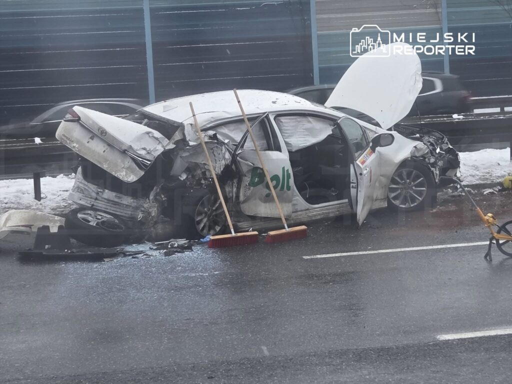 Zderzony samochód osobowy z widocznymi uszkodzeniami na drodze, obok leżą miotły i fragmenty szkła.