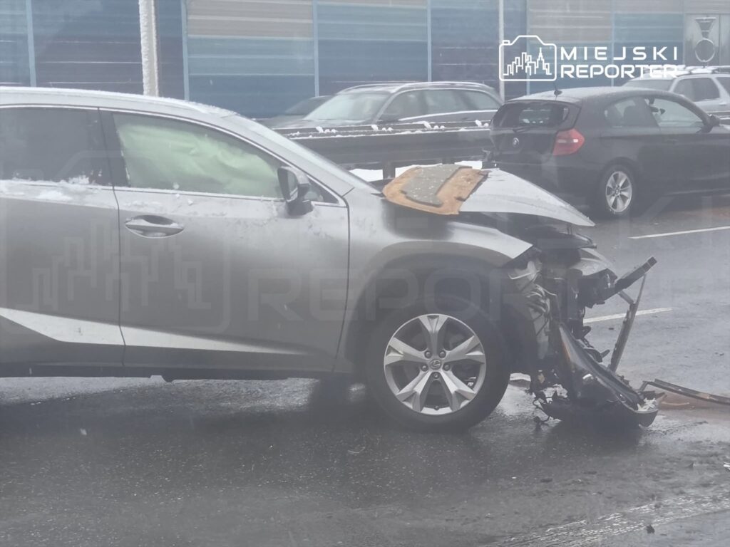 Zniszczony samochód osobowy z uszkodzoną przednią częścią, zaparkowany na mokrej jezdni w deszczowej aurze.