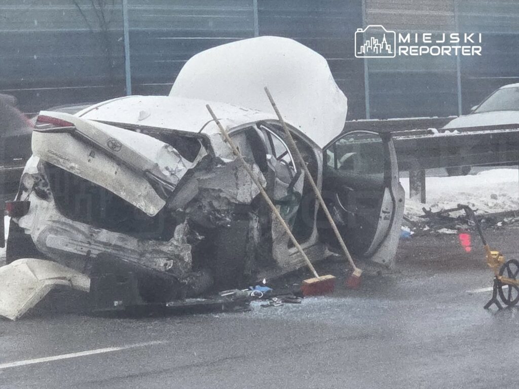 Uszkodzony biały samochód osobowy leży na boku na drodze, z otwartymi drzwiami i rozbitym przodem. Obok znajdują się miotły i fragmenty szkła.