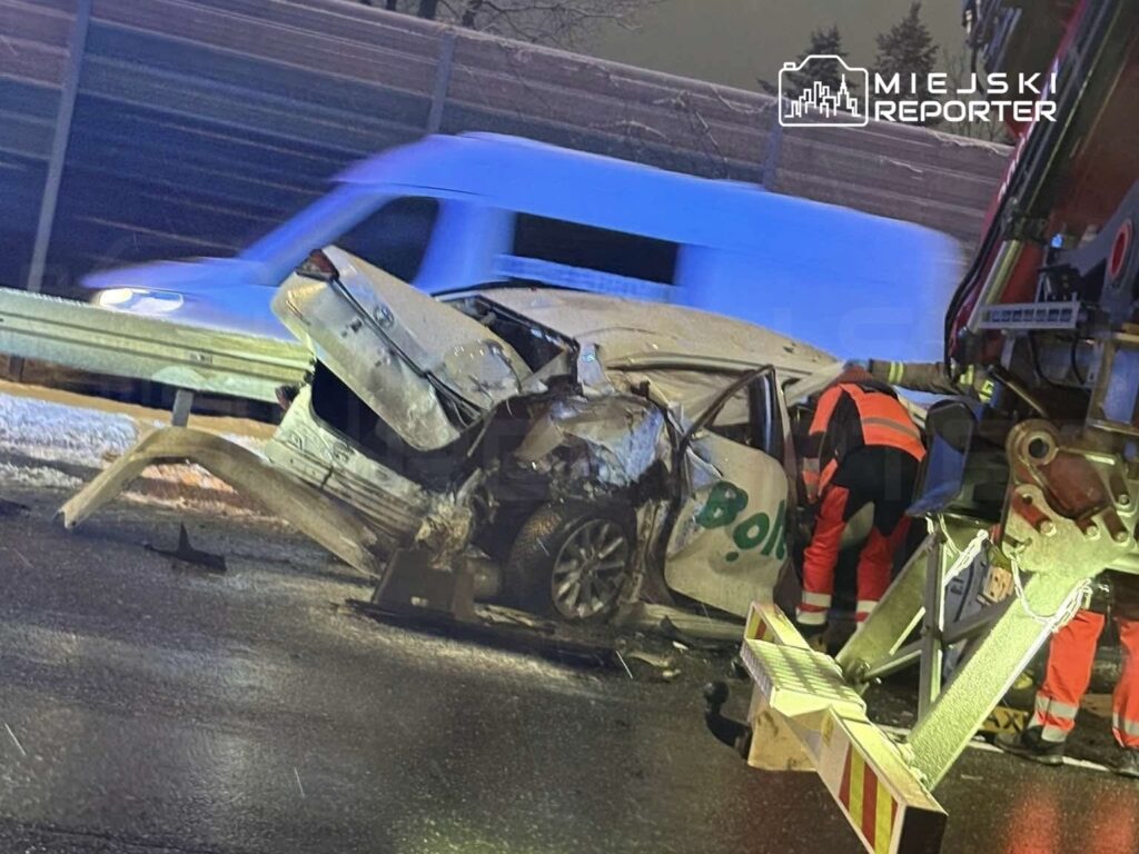 Rozbity samochód osobowy leży na jezdni, obok interweniujący ratownicy w odblaskowych strojach.