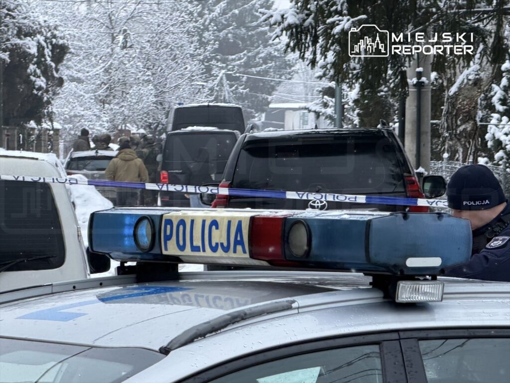 Policyjny radiowóz z włączonymi sygnalizatorami na zaśnieżonej ulicy, w tle grupa funkcjonariuszy i pojazdów.