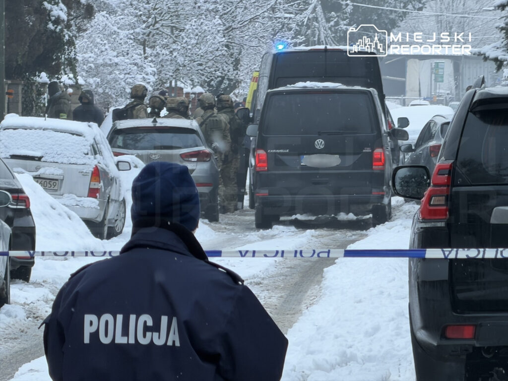 Policjant w niebieskiej czapce obserwuje działania służb mundurowych na zaśnieżonej ulicy.