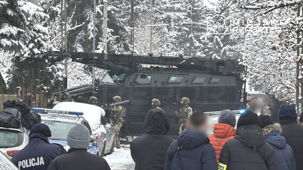 Grupa uzbrojonych żołnierzy stoi w pobliżu opancerzonego pojazdu na zaśnieżonej ulicy, obserwując otoczenie.